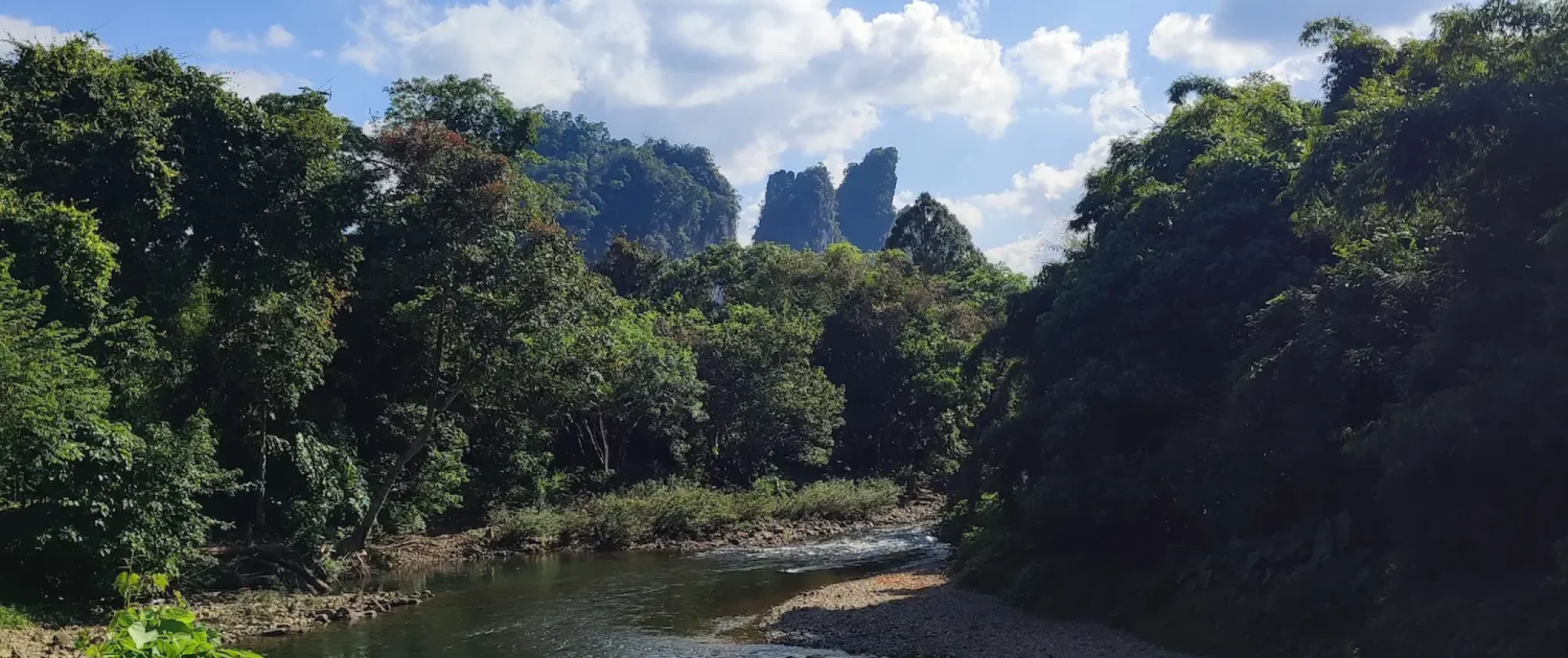 Kavicsos medrű Sok folyó sűrű esőerdő mellett, a háttérben a Khao Sok Nemzeti Park ikonikus mészkőszirtjeivel a kék ég alatt.