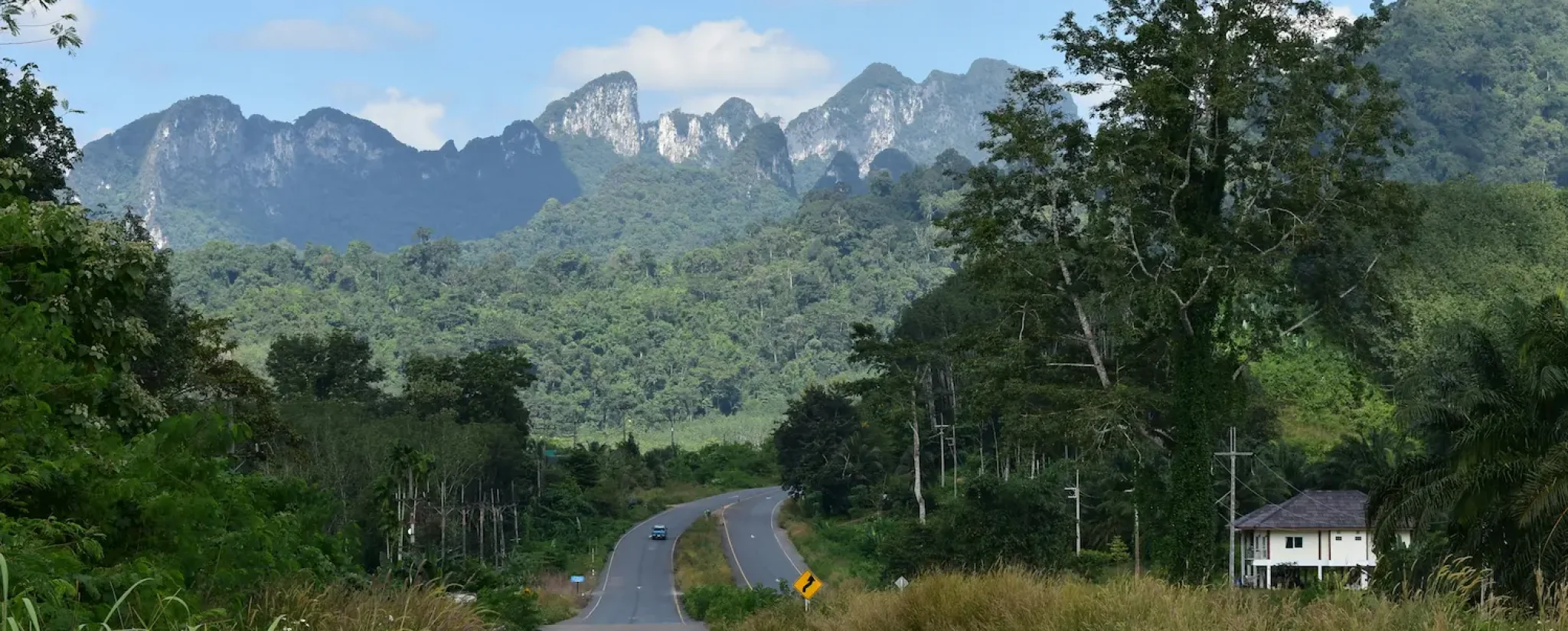 Kanyargós aszfaltút vezet a monumentális, zöld növényzettel borított mészkősziklák felé a thaiföldi Khao Sok Nemzeti Parkban, kék égbolttal és fehér felhőkkel – a Dream Journey szóló utazás transzformatív megérkezésének képe.